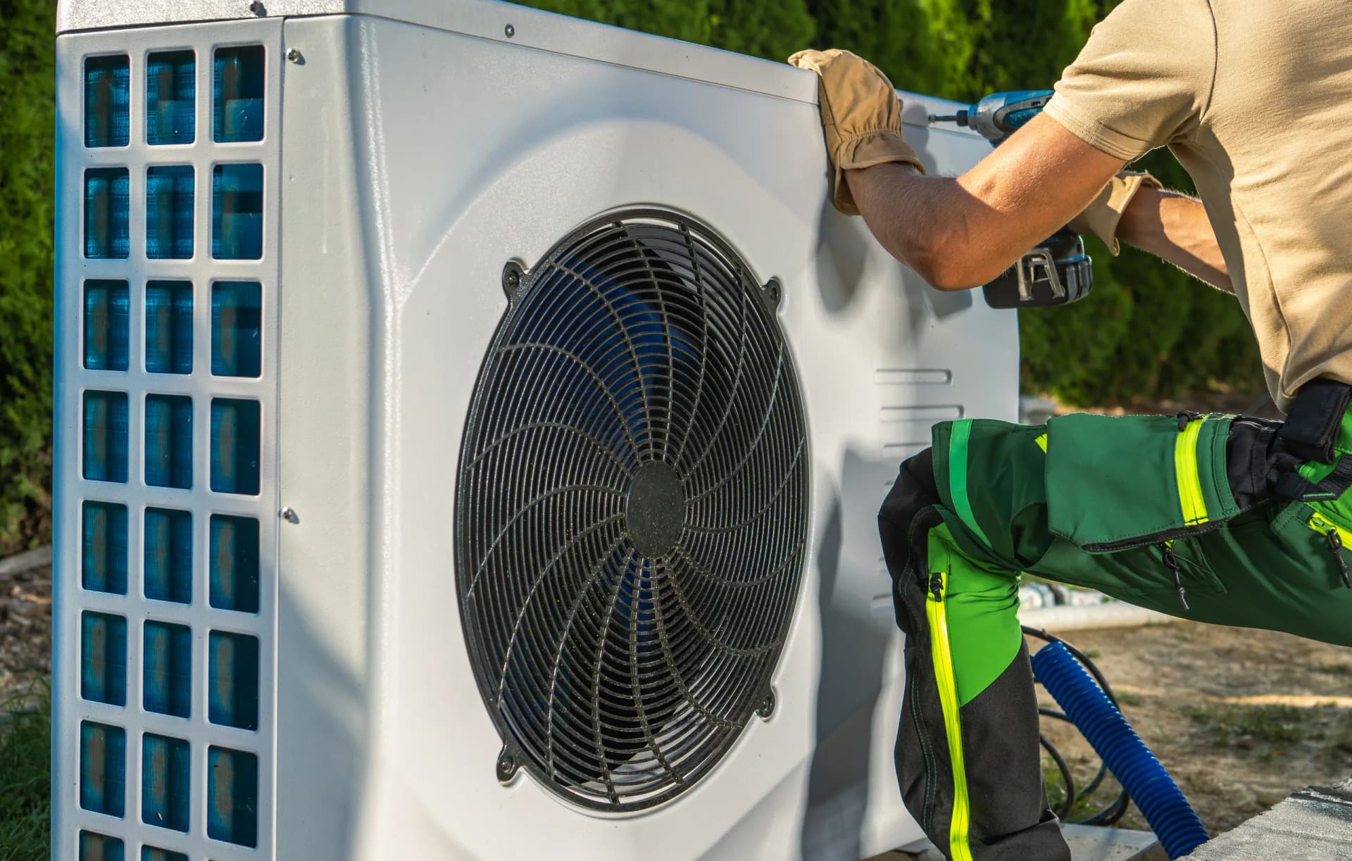 A heating engineer works on an outdoor air source heat pump or air conditioning unit with a power drill.
